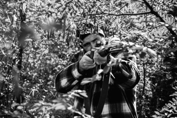 Person in a plaid shirt aiming a firearm in a forest setting.