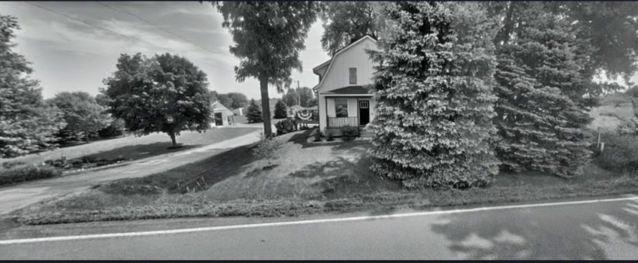 Black and white photograph of a house surrounded by trees and a road.