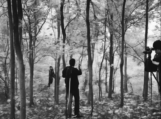 Three figures in dark clothing stand among trees in a foggy forest setting.